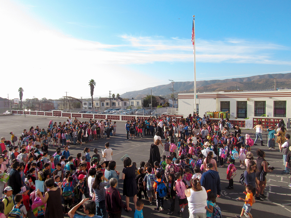 VVES Morning Circle - The whole school of students are standing in a circle, in front of the flag on the yard.