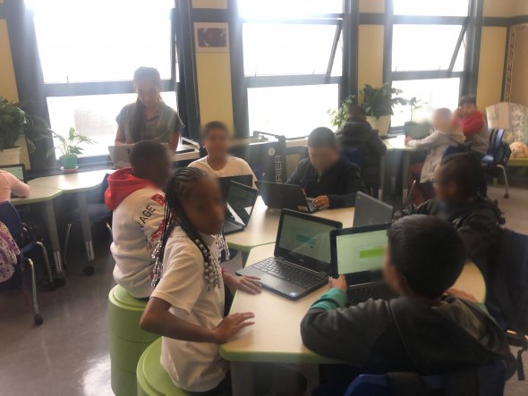 Students working in groups at flexible seating desks, with laptops in front of them, and a teacher in the background