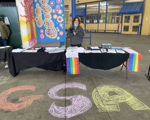 Zachary Lipsett, an LGBTQ+ youth activist and recent graduate from Ruth Asawa School of the Arts (SOTA), stands at a table in front of a GSA sidewalk chalk sign.