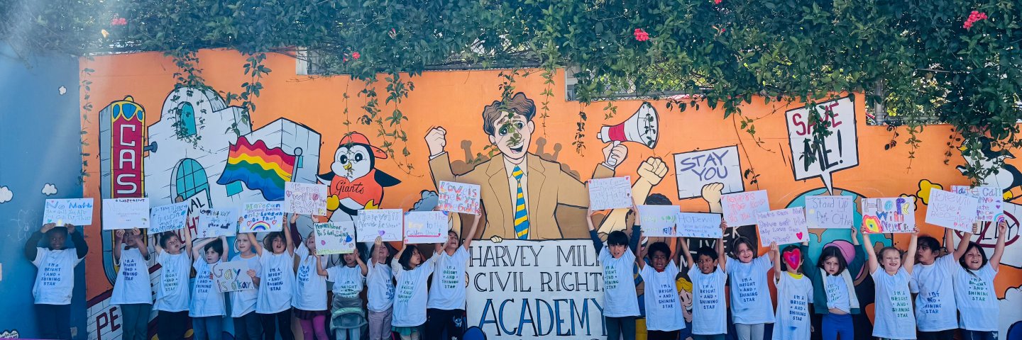 First grade students stand with handmade signs in front of a colorful mural with an illustration of Harvey Milk and other characters and Castro landmarks.