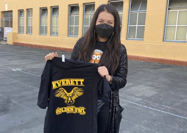 Esther Fensel, Principal at Everett Middle School, wears a mask and holds up an Everett t-shirt