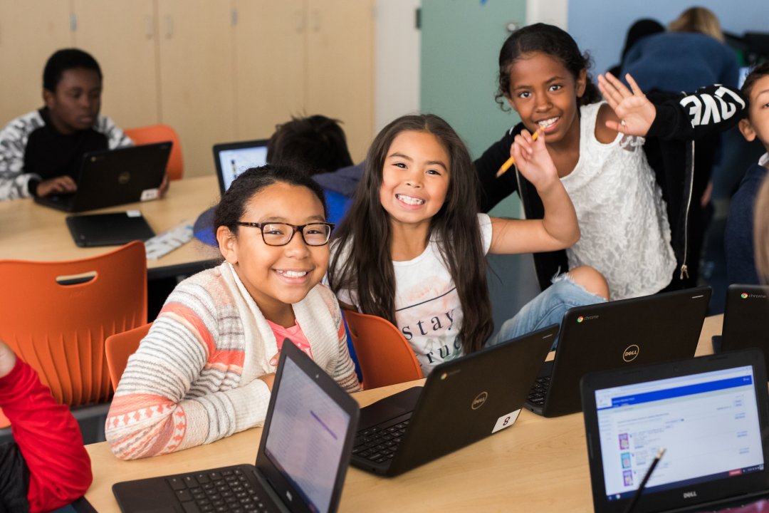 Three students, one with glasses, one with a pencil in hand, and another waving, work on their laptops and smile at the camera.