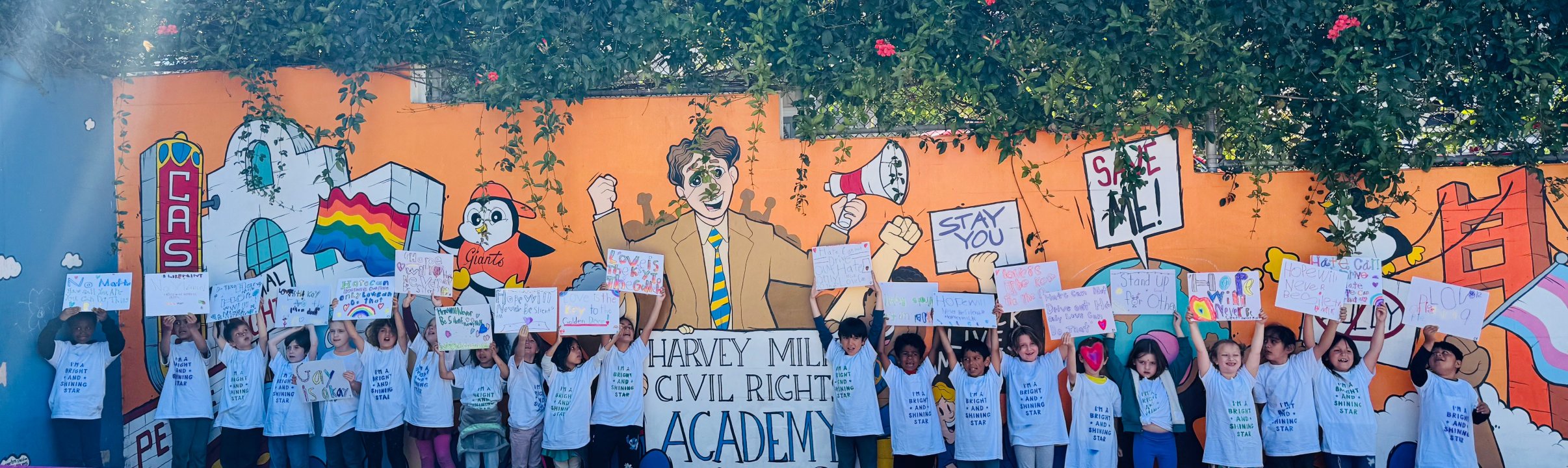 First grade students stand with handmade signs in front of a colorful mural with an illustration of Harvey Milk and other characters and Castro landmarks.
