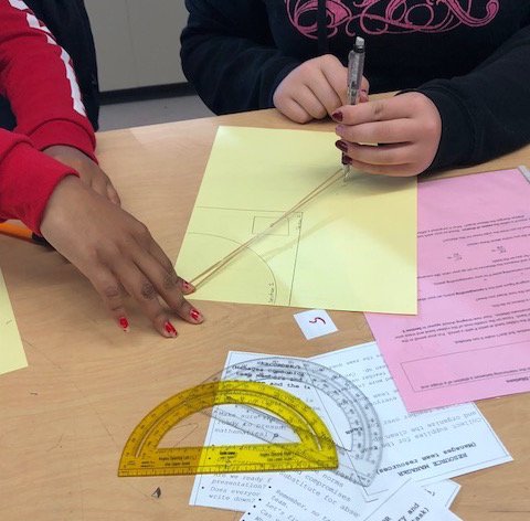 Hands of two students using a rubberband to enlarge an image. Protractor and geometry papers on the desk.
