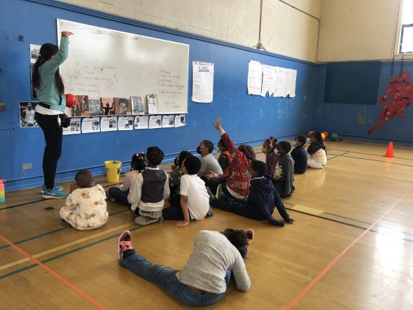 Students sitting as a group in a gym receiving instructions from their teacher.