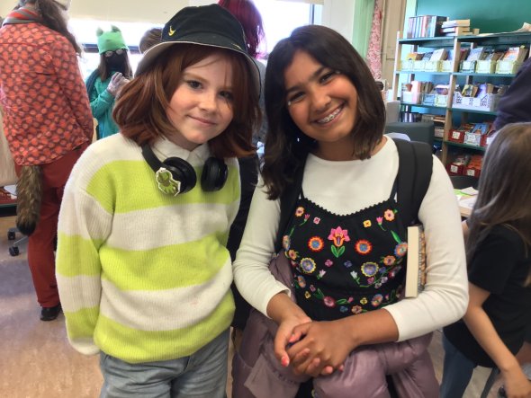 Two 5th-grade students with neck-length hair, one redhead and one brunette, standing side by side and smiling