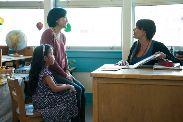 Image of a classroom with a young woman at a desk, holding a binder open and speaking to a middle aged woman and young girl who are seated next to her.