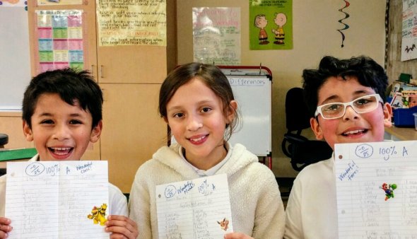 Three elementary students smile in a classroom holding up their test papers.