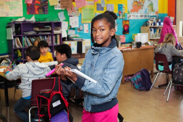 Picture of an African American girl in a classroom, holding a notebook and smiling