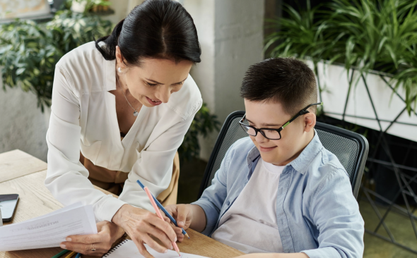 A woman with dark hair and a white blouse is helping a boy with short dart hair and glasses with papers on a desk