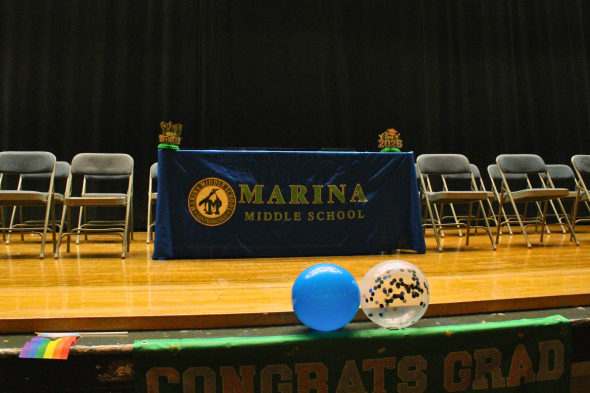 there is a table on the stage with a marina middle school tablecloth including the school logo with 2 chairs on the left and 2 chairs on the left. in front of the stage, there is a pride flag laying down on the left side of the screen and 2 balloons (one blue and one white with glitter) inside right in front of the table. below the pride flag and balloons is a banner that says "congrats grad" that is cut off. 