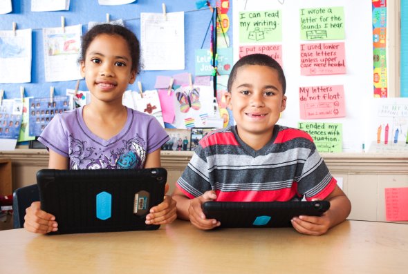 A boy and a girl are smiling while holding their tablets.