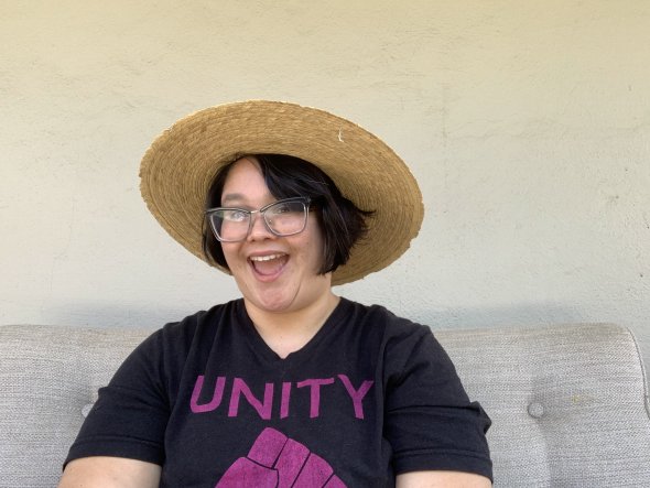 a picture of a mixed raced woman wearing a grey t-shirt that says "UNITY", a straw summer hat, and a goofy grin