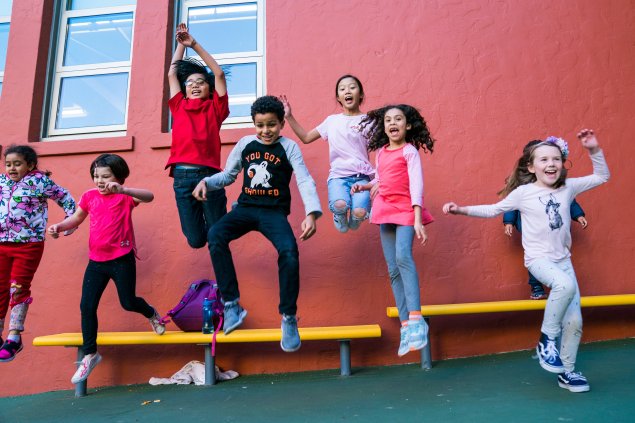 Seven students jumping in the air off a yellow bench smiling in front of a red wall. 