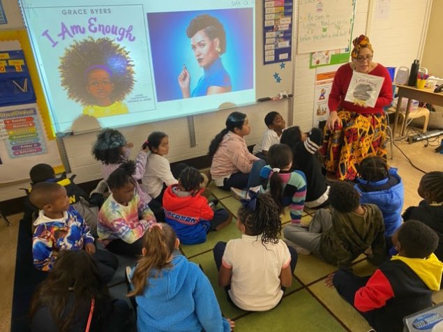 Students seated look at educator holding a book showing the cover