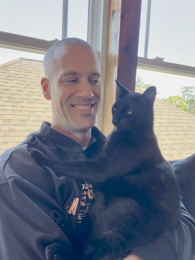 Male individual smiling while holding black cat in arms