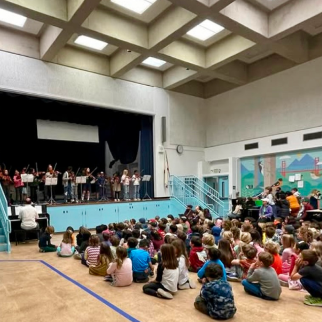 An image showing a large group of elementary students sitting on the floor of a high-ceilinged auditorium, watching a group of their peers perform on stage. Approximately ten fourth and fifth-grade students stand on a raised stage behind music stands, playing violins while an adult accompanies them on a piano to the left. The auditorium features a distinctive coffered ceiling with skylights and a large colorful mural on the right wall depicting San Francisco landmarks like the Golden Gate Bridge.