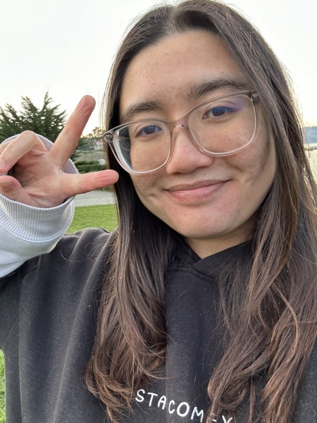 A person with long brown hair and glasses flashes a peace sign and smiles at the camera