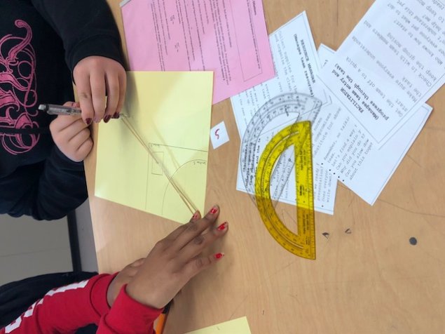 Hands of two students using a rubberband to enlarge an image. Protractor and geometry papers on the desk.