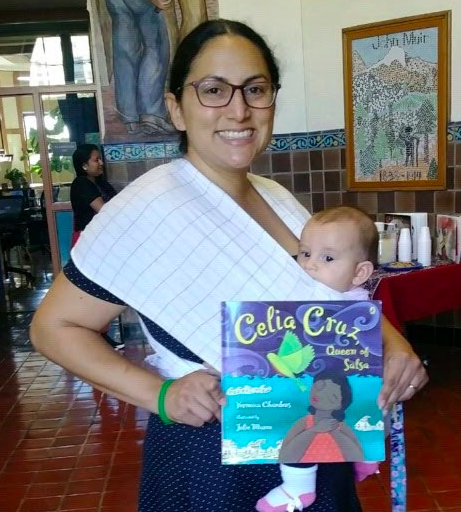 Nercy Arias smiling holding her baby and a picture book about Celia Cruz